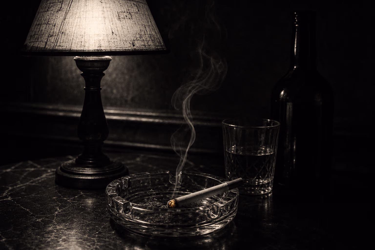 Noir-Stillleben im Dunkeln: eine Tischlampe, eine glimmende Zigarette im Aschenbecher mit bernsteinfarbener Glut sowie Glas und Flasche.
