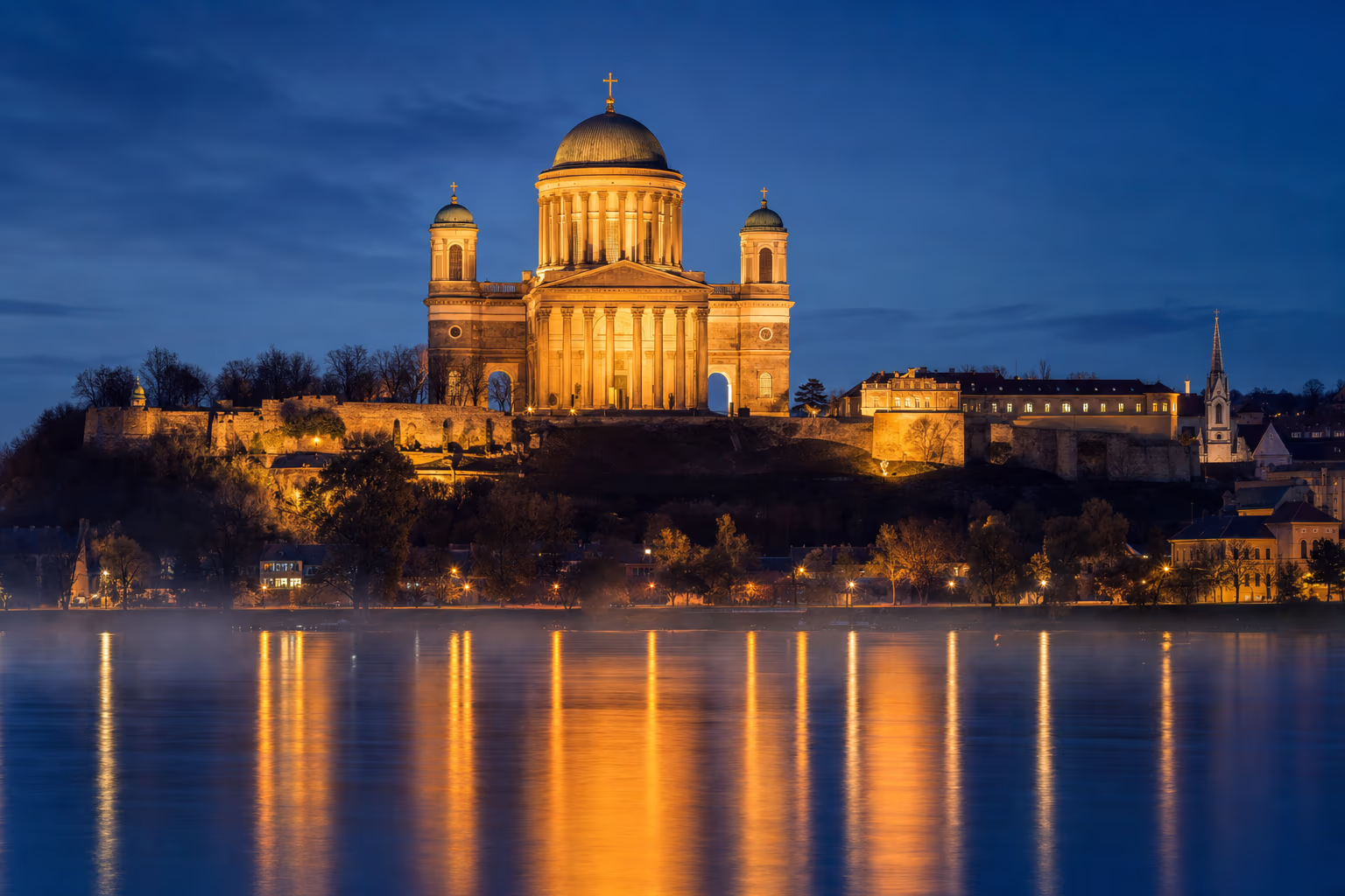Die Basilika von Esztergom zur blauen Stunde, von der Donau aus gesehen: die gold beleuchtete Kuppel auf dem Burgberg, mit langen goldenen Spiegelungen auf dem dunklen Fluss (KI-Illustration).