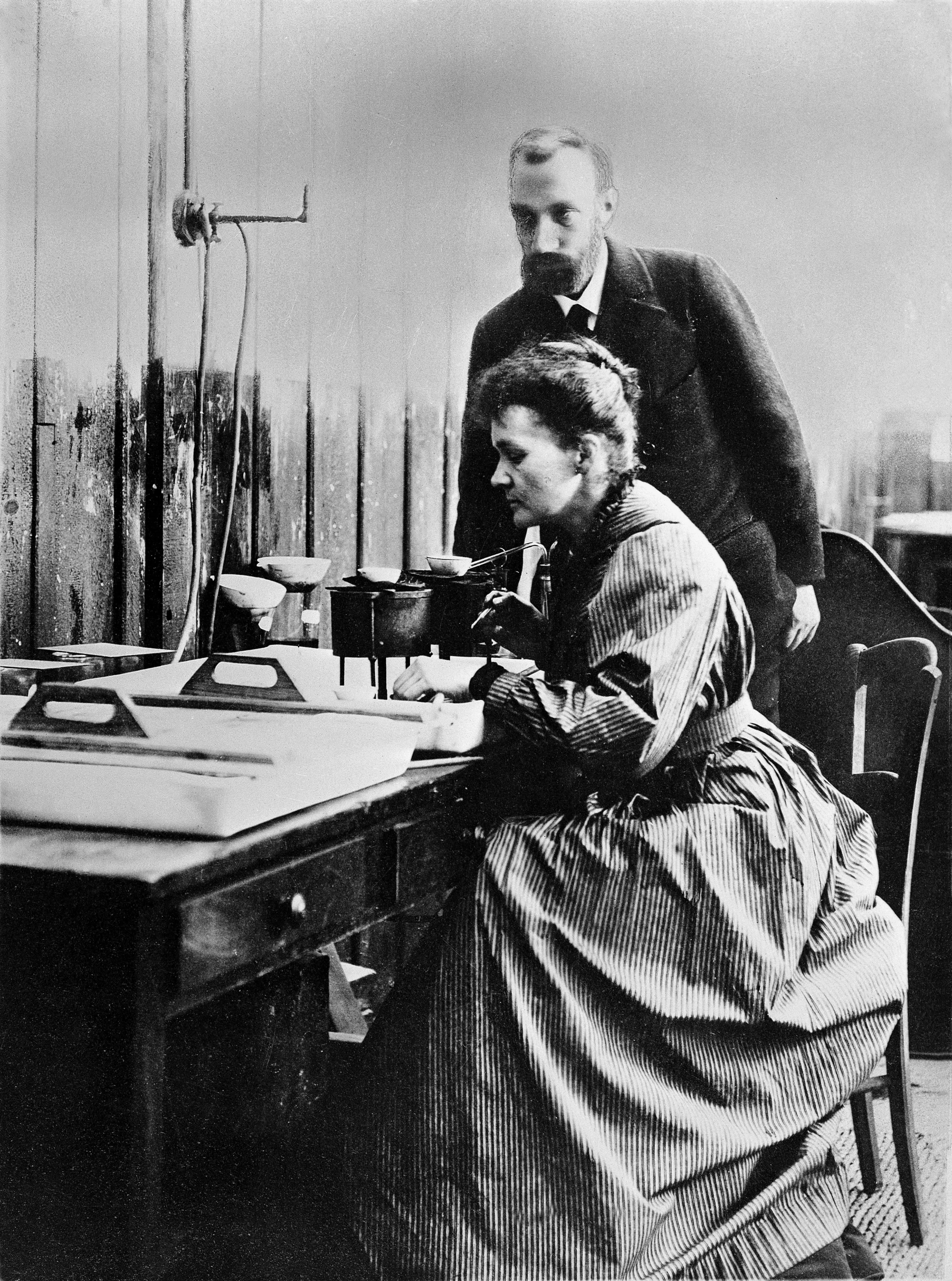 Historische Fotografie: Marie Curie sitzt an einem Labortisch mit Messapparatur, Pierre Curie steht hinter ihr, im einfachen Pariser Labor